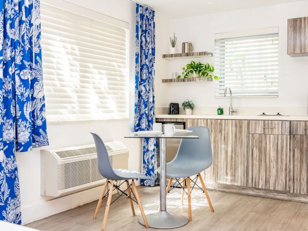 A bright kitchen-dining area with blue patterned curtains, a small round table, two light blue chairs, and wooden cabinets with a countertop.