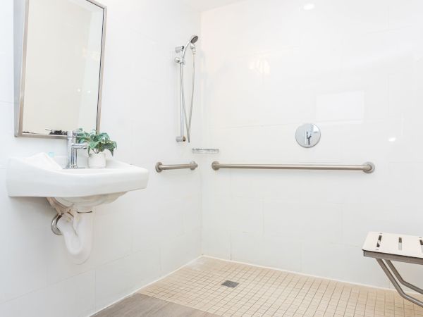 A bright, accessible bathroom with a wall-mounted sink, mirror, and grab bars; a shower area with a handheld spray and a small stool.