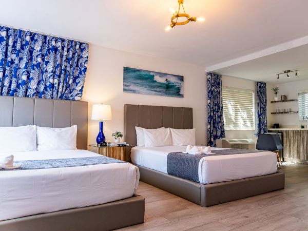 A bright hotel room with two large beds, blue patterned drapes, a bedside lamp, seaside artwork, and a small sitting area with a kitchenette in the background.
