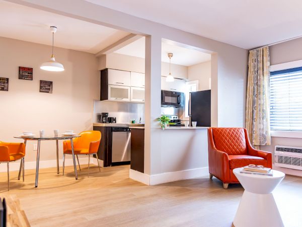 A bright, modern apartment with an open-plan kitchen and dining area; orange chairs, a red armchair, wooden floors, and a large window with blinds.