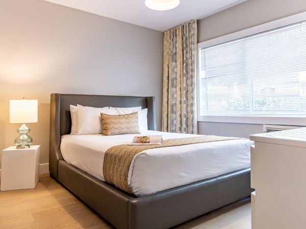 A modern hotel room with a plush bed, two pillows, bedside lamp, white nightstand, and a window with curtains.