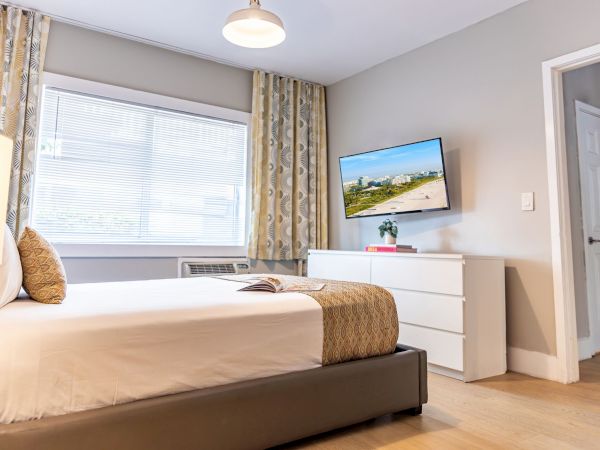 A tidy hotel-style bedroom with a queen bed, white dresser, wall-mounted TV, large window with patterned curtains, and soft modern decor.