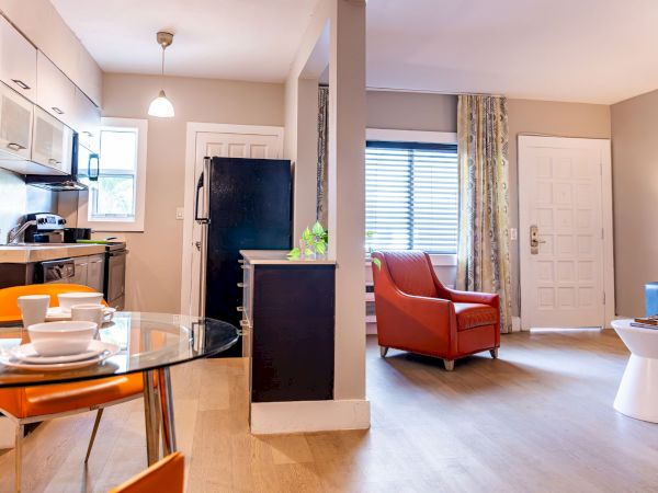 A bright open-plan apartment with a compact kitchen, dining area with orange chairs, and a living room featuring a red chair, white side table, and a blue couch.