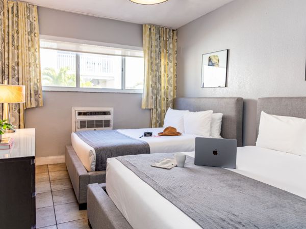 Two tidy hotel rooms side by side with two beds, a laptop on the near bed, a dresser, window with curtains, and framed art on the walls.