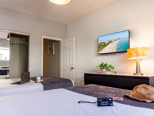 A bright hotel room with two beds, a dresser, wall-mounted TV, plant, lamp, hat and sunglasses on the bed, and an open doorway to a small closet or bathroom.