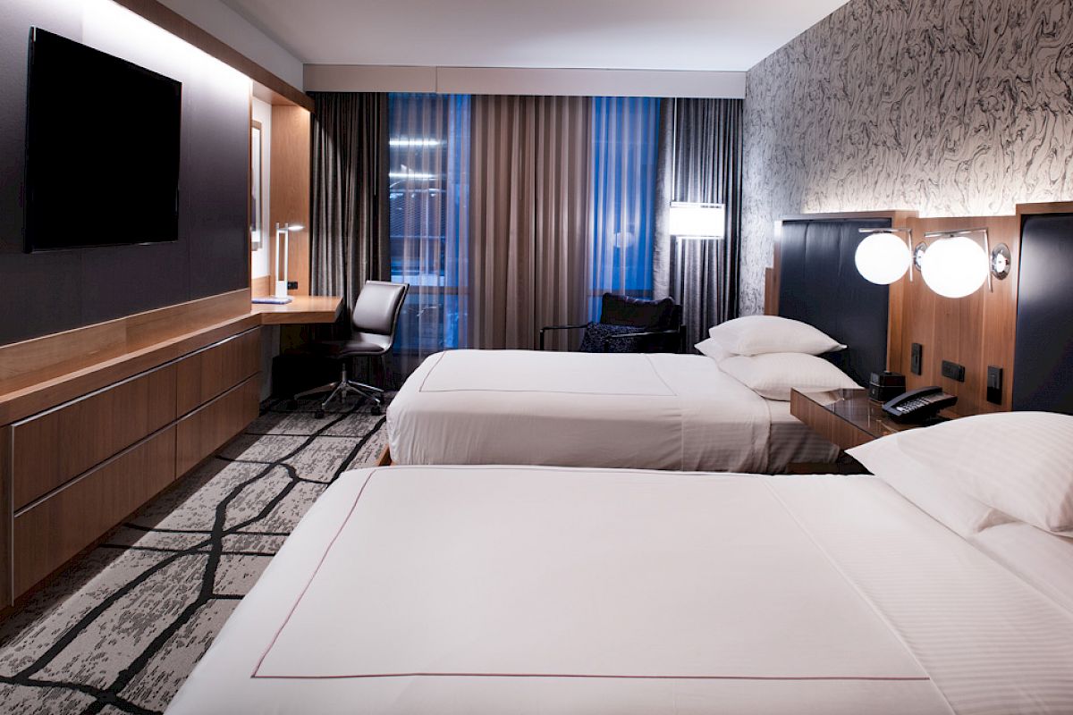 A modern hotel room features two beds, a TV, a desk with a chair, and stylish decor.