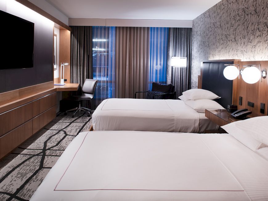 A modern hotel room features two beds, a TV, a desk with a chair, and stylish decor.