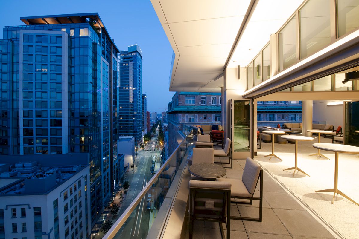 Modern rooftop terrace with seating overlooks a city street and high-rise buildings at dusk, capturing an urban evening scene.