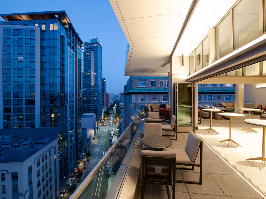 Modern rooftop terrace with seating overlooks a city street and high-rise buildings at dusk, capturing an urban evening scene.