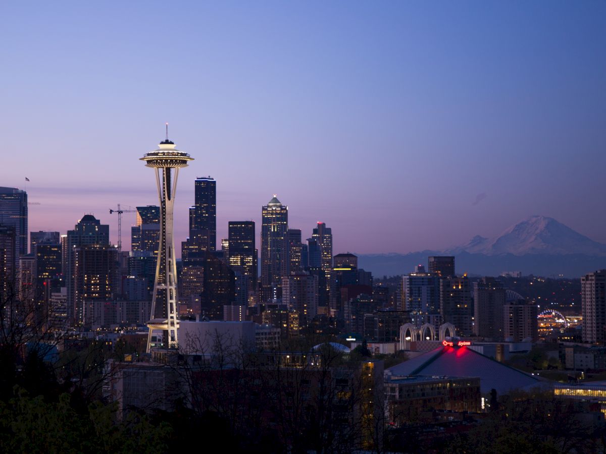 Seattle skyline at dusk with the Space Needle prominently visible against the cityscape and a mountain in the background.