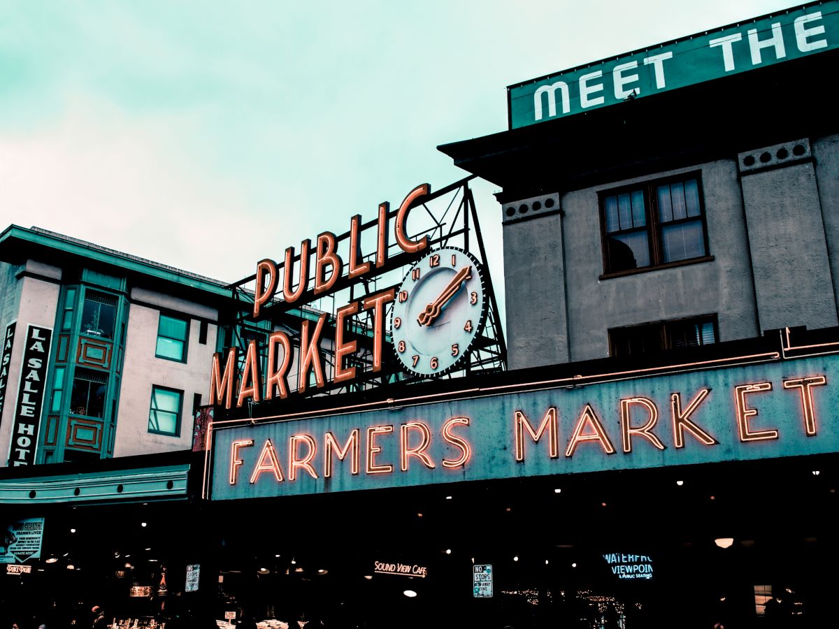 The image shows the iconic signage of a public market with a clock and neon lights, promoting a farmers market.