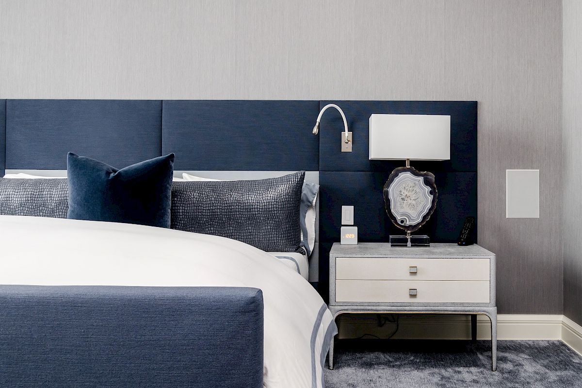 A modern bedroom with a navy blue headboard, white bedding, and a nightstand featuring a lamp and decorative item.