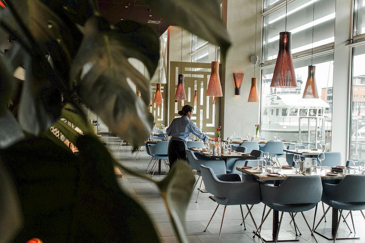 A modern restaurant interior with a person setting tables, stylish pendant lights, and green plants in the foreground complete the scene.