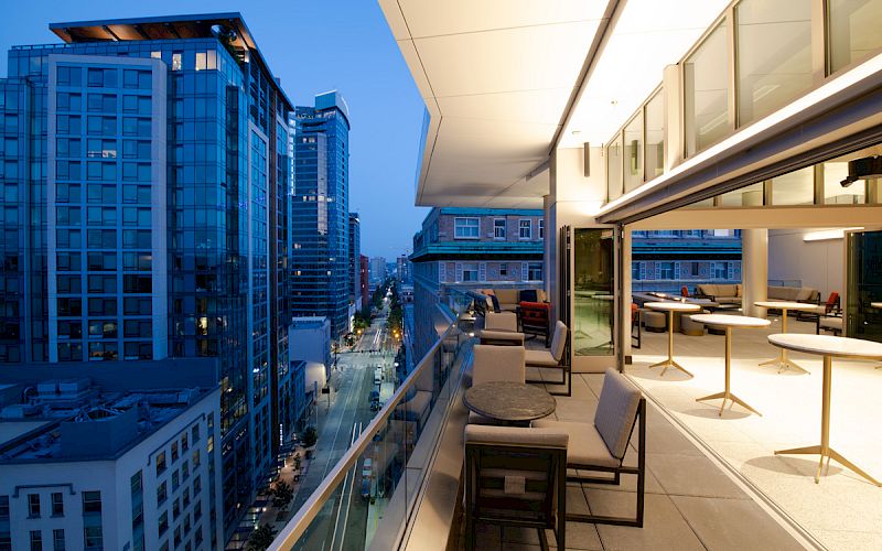 A modern cityscape at twilight, with tall buildings and a stylish glass-walled balcony featuring tables and chairs.
